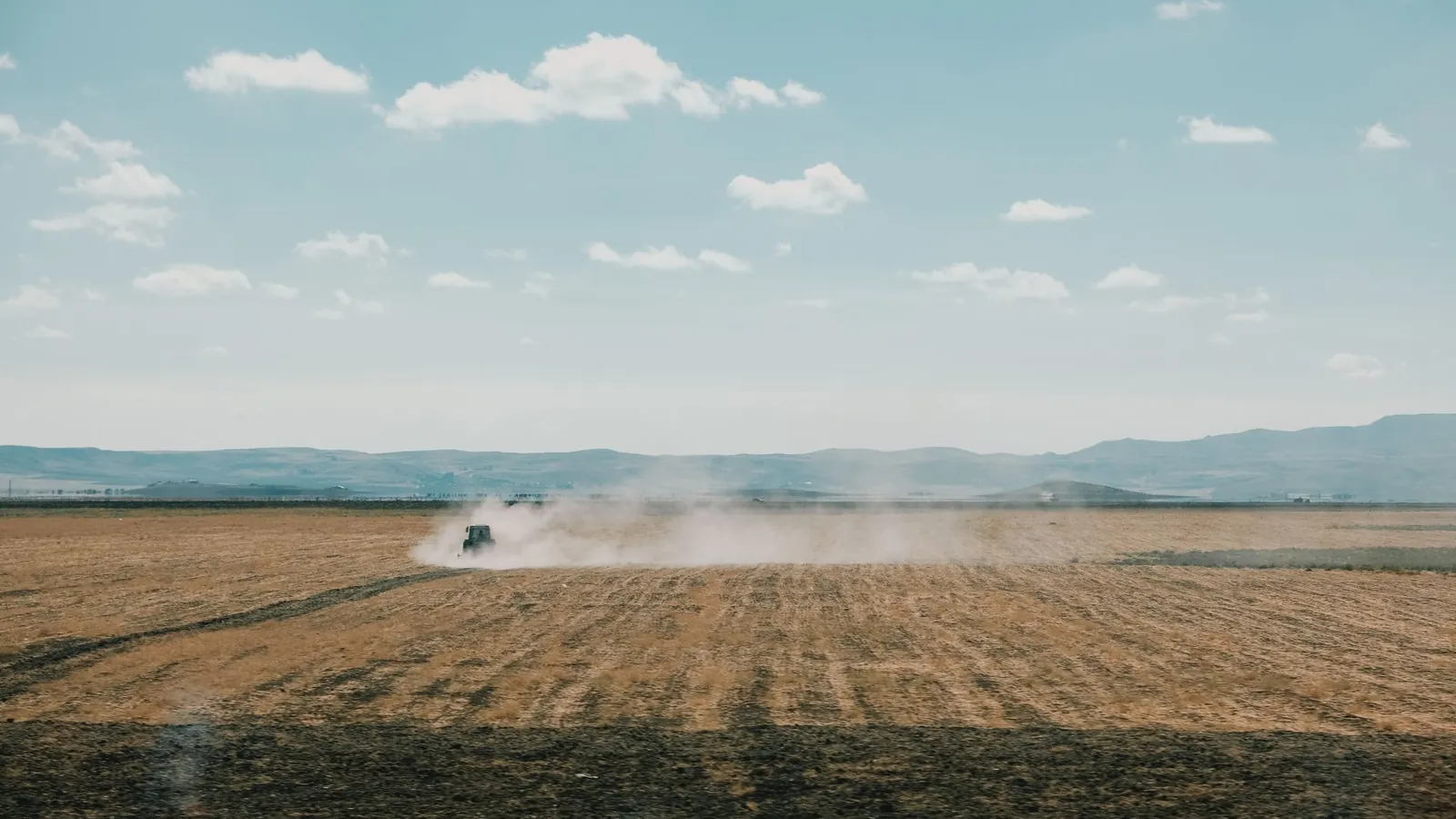 Tracteur traversant une vaste parcelle moissonnée en soulevant un long nuage de poussière